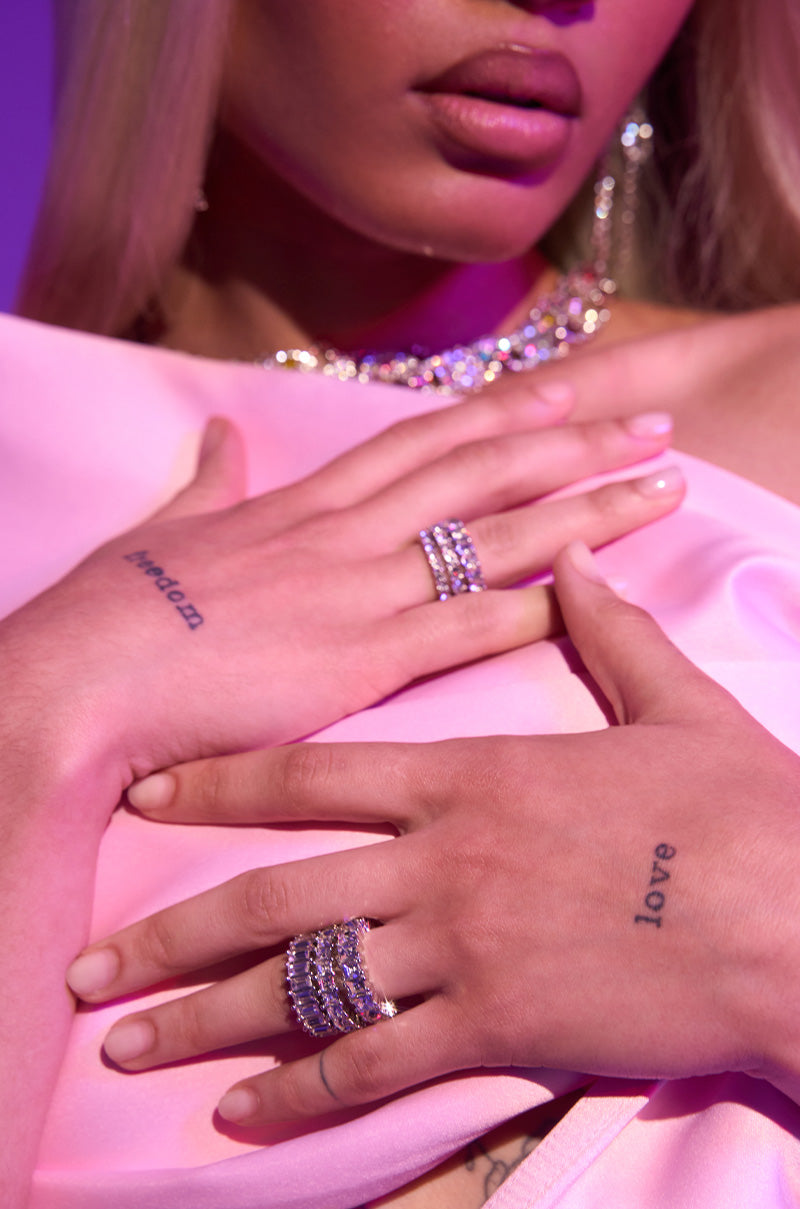 A close-up of someone wearing the SPEND IT RHINESTONE RING SET and silver-tone bands, plus a jeweled necklace. "LOVE" and "LOVED" tattoos on their hands, in soft pink fabric under purple lighting.