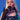 A person with long platinum blonde hair poses against a blue backdrop in an AKIRA Label dress with mirror shards, black mesh gloves, YUCK RINGS in faux silver, and a jeweled cross necklace, covering their mouth with a gloved hand.
