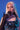 A person with long platinum blonde hair poses against a blue backdrop in an AKIRA Label dress with mirror shards, black mesh gloves, YUCK RINGS in faux silver, and a jeweled cross necklace, covering their mouth with a gloved hand.