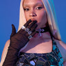 A person with long platinum blonde hair poses against a blue backdrop in an AKIRA Label dress with mirror shards, black mesh gloves, YUCK RINGS in faux silver, and a jeweled cross necklace, covering their mouth with a gloved hand.