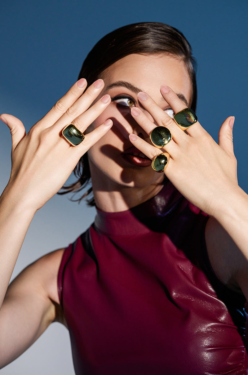 A woman in a sleeveless burgundy top poses with her hands covering part of her face, highlighting the TREND DRIVEN RING SET—large gold rings with dark green stones—against a blue and white background.