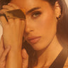 A woman with long brown hair gazes at the camera, her hand to her face as she showcases the MOD LIFE RING—a bold, large gold geometric statement piece—paired with black earrings and illuminated by warm, soft lighting.
