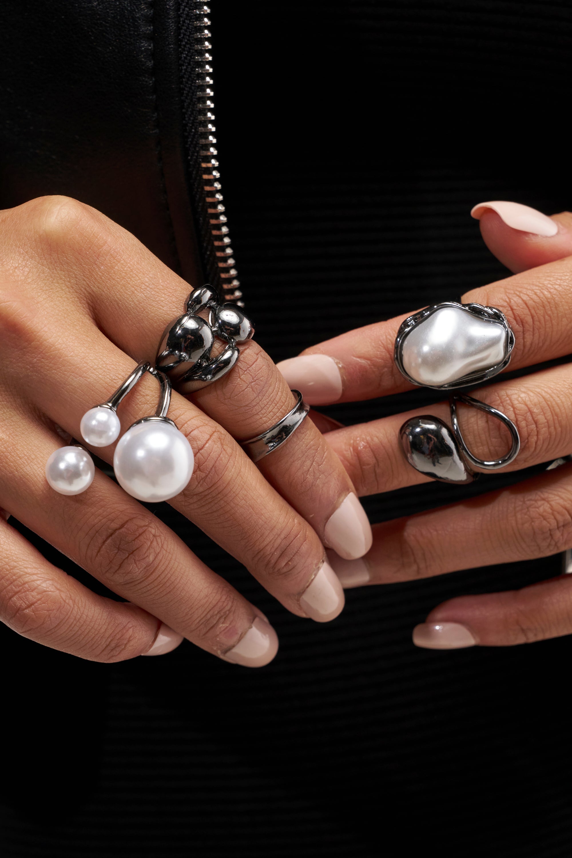Close-up of hands with manicured nails wearing chunky chrome metallic rings and the CHROME CRUSH PEARL RING SET, adorned with large white pearls, against a black zippered jacket.