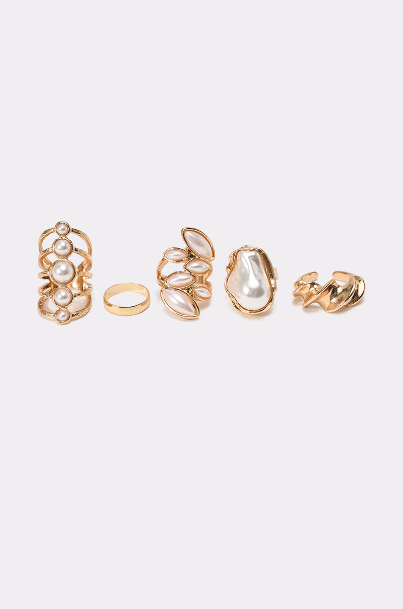 The VANJA RING SET features five gold-tone rings with faux pearl accents, oval stones, and abstract shapes. Each ring has unique detailing and is displayed in a row against a plain light background. 2