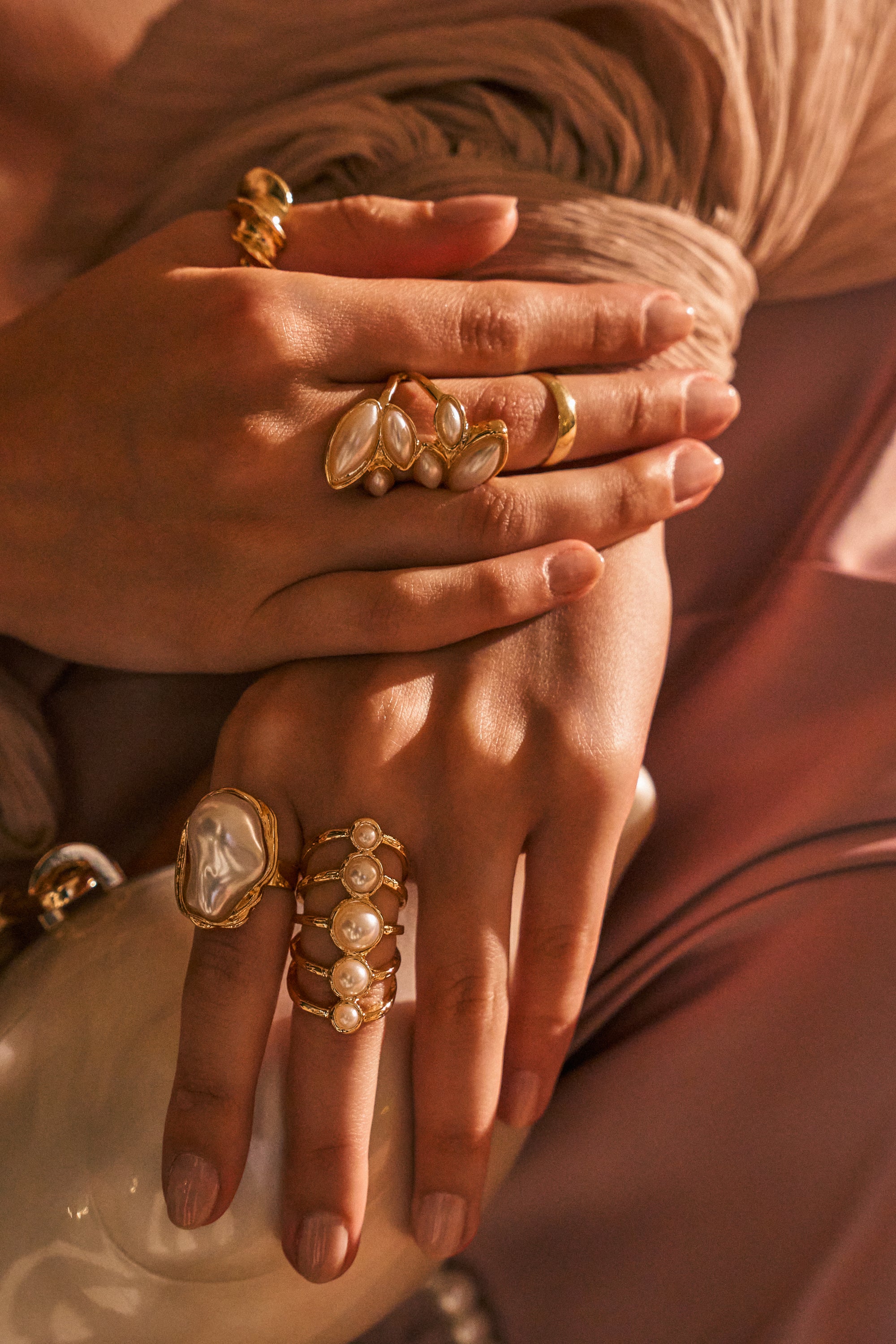 Two hands adorned with multiple VANJA RING SET rings are shown resting on a shiny, curved white object, with soft beige and pink fabric in the background.