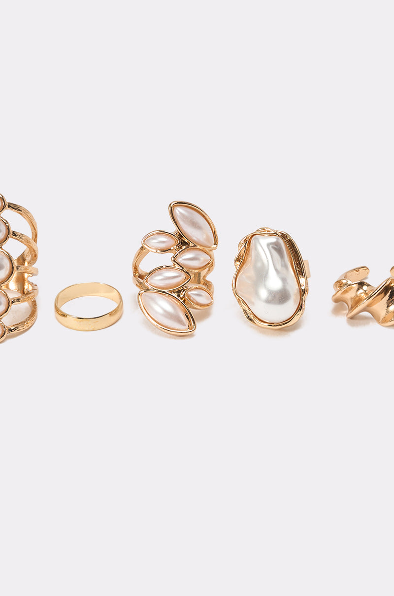 The VANJA RING SET features five vintage-inspired gold-tone stackable rings with intricate designs and white pearls, displayed in a row on a light gray background. 4