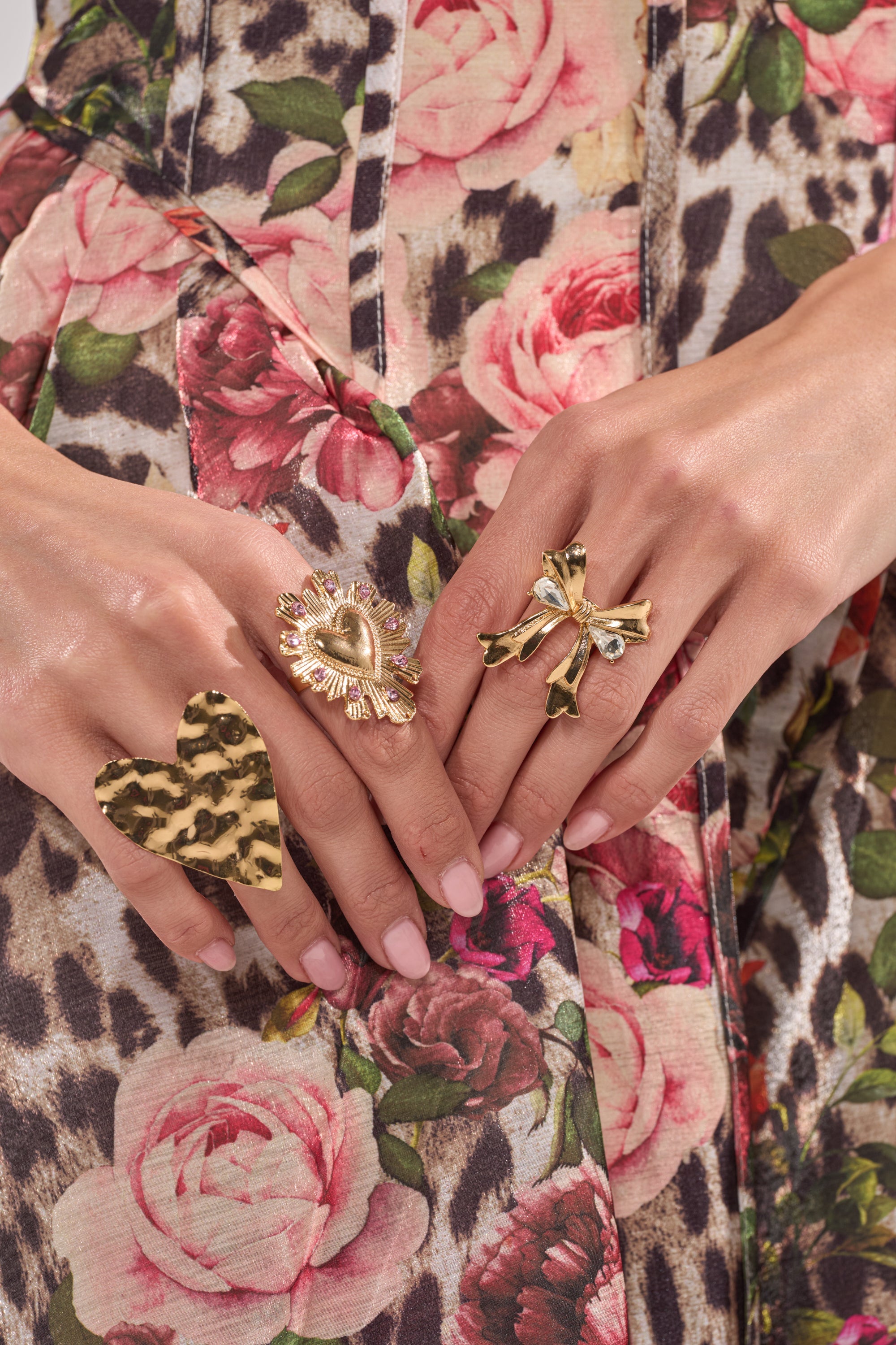 Wearing the CROSS MY HEART RING SET, which features an adjustable leopard print heart ring, a gold heart ring with rhinestones, and a gold bow ring, someone shows off manicured nails against a floral and animal print fabric. 0