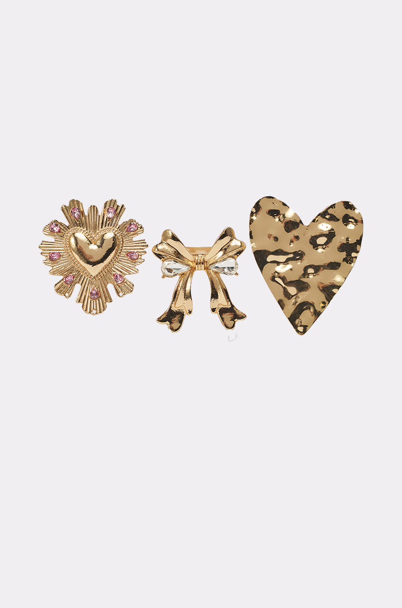 The CROSS MY HEART RING SET features three metallic rings: a gold heart on sunburst with pink accents, a gold and silver bow with rhinestones, and a hammered gold heart with leopard print, shown on a plain white background. 1