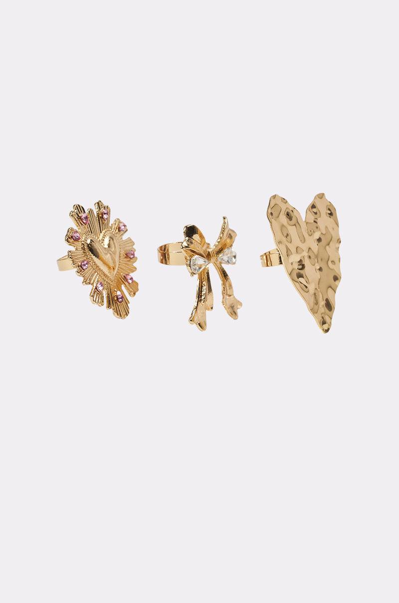 The CROSS MY HEART RING SET includes three gold-tone rings: one with a textured heart and pink accents, one styled as a crystal bow, and one featuring a large hammered heart. 2