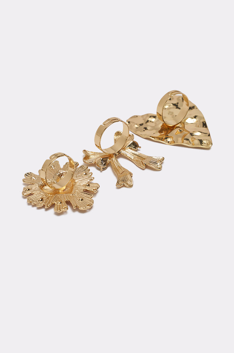 The CROSS MY HEART RING SET includes three gold-tone adjustable rings—a flower, a bow, and a textured heart—displayed on a white background. 3