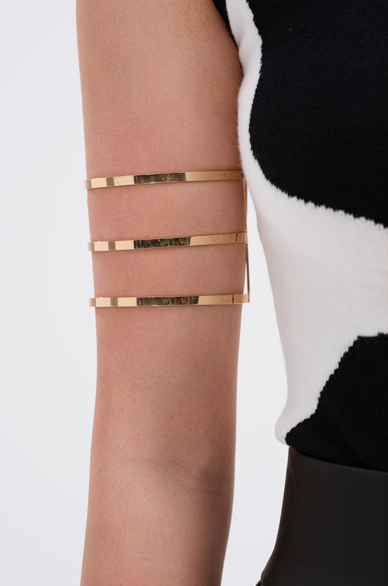 A close-up of a person’s upper arm accessorizing with three STRICTLY BUSINESS ARM CUFFS in faux gold, paired with part of a black and white sleeveless top. 1