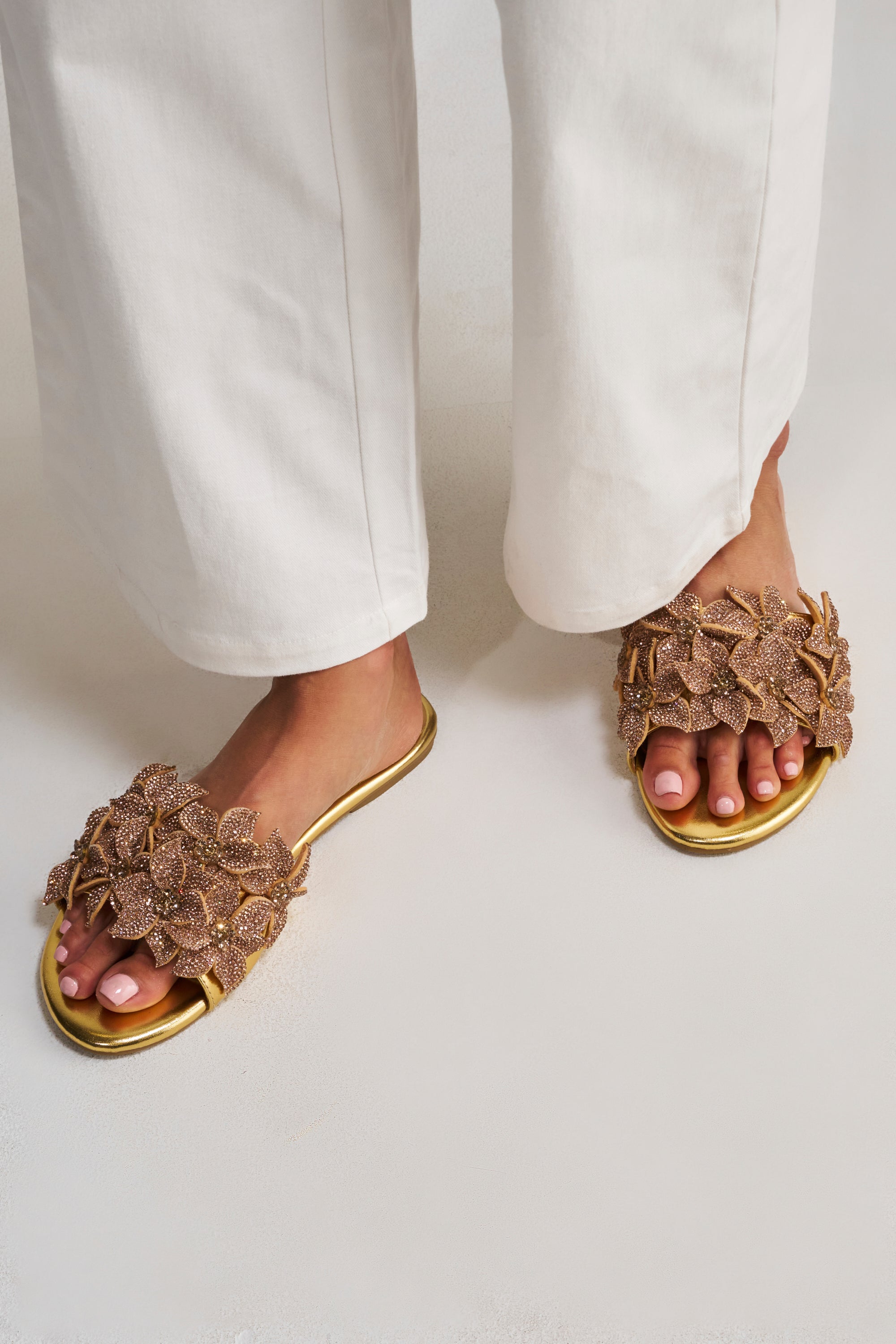 A person wearing white pants and AZALEA WANG YUNA Gold Flat Rhinestone Embellished Slip On Sandals stands on a light-colored surface.