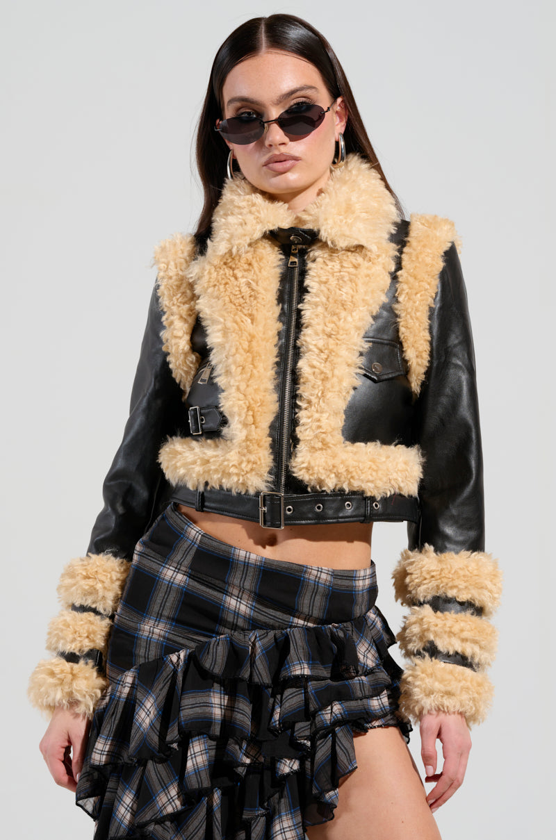 A person wearing sunglasses showcases the YURI SHERPA MOTO JACKET with black and tan fur trim, paired with a plaid ruffled skirt. Their long dark hair complements their confident pose against a light gray background.