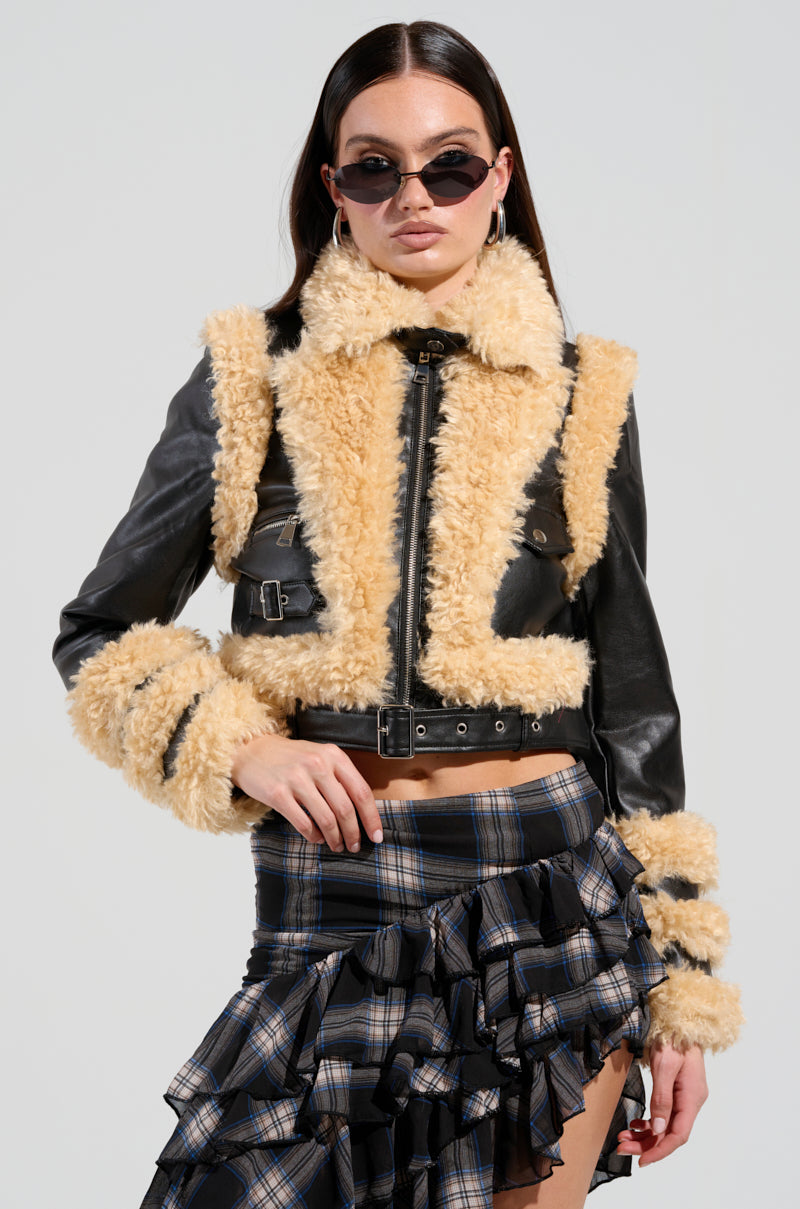 A woman is wearing the chic YURI SHERPA MOTO JACKET in black and tan with buckle details, paired with a plaid ruffled skirt. Her long dark hair flows behind oval sunglasses as she stands against a plain background, exuding effortless chic.