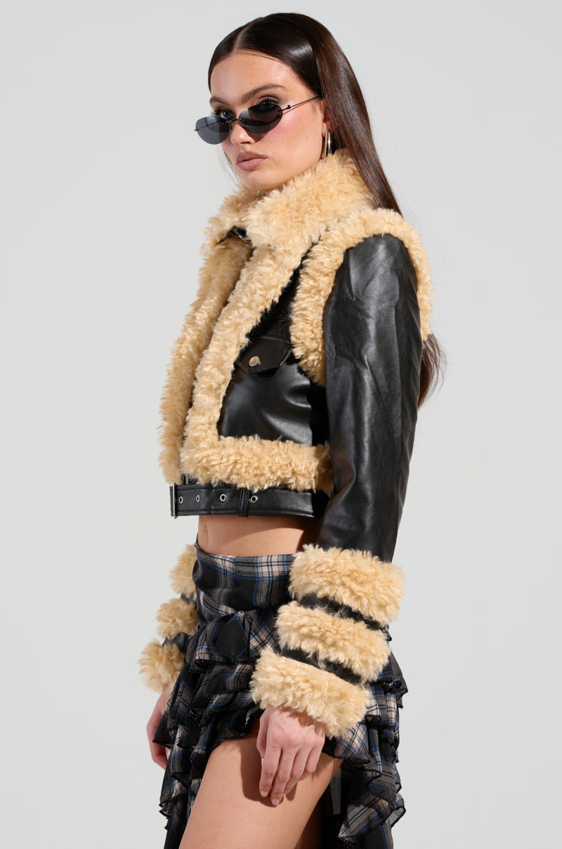 The person is wearing the YURI SHERPA MOTO JACKET, which is a black vegan leather jacket with beige faux sherpa trim, along with a plaid skirt and black sunglasses. The moto jacket includes fur details on the collar, cuffs, and hemline. They have long brown hair and are standing against a plain background.