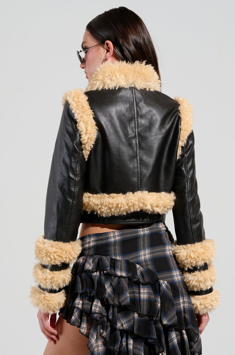 A person with long dark hair, wearing sunglasses, is seen from behind. They are dressed in a YURI SHERPA MOTO JACKET and a plaid, ruffled skirt.