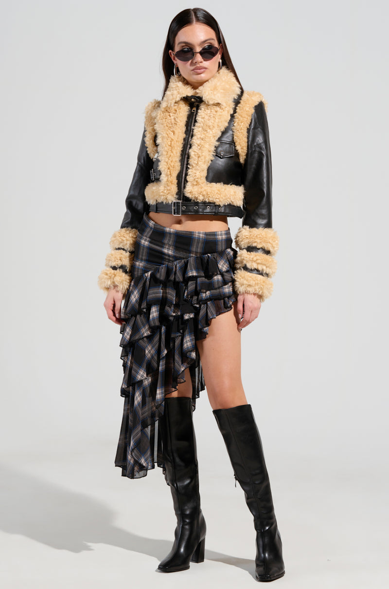 A woman in sunglasses wearing the YURI SHERPA MOTO JACKET, featuring black vegan leather with shearling details, paired with an asymmetrical plaid skirt and knee-high black boots, stands against a plain background.