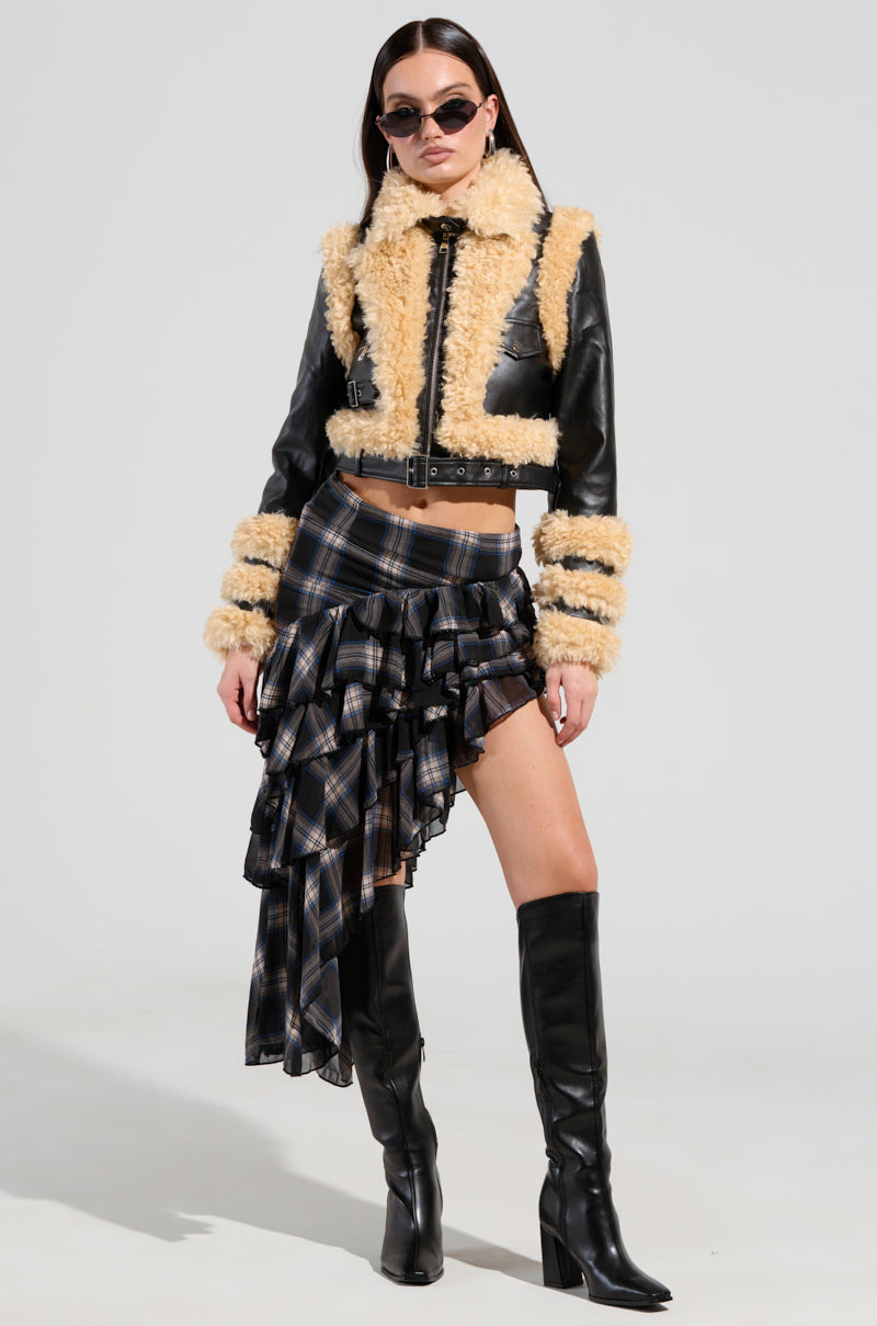 A woman wearing sunglasses and the YURI SHERPA MOTO JACKET over a plaid asymmetrical skirt with layered ruffles, paired with knee-high black boots. She stands confidently against a plain background.