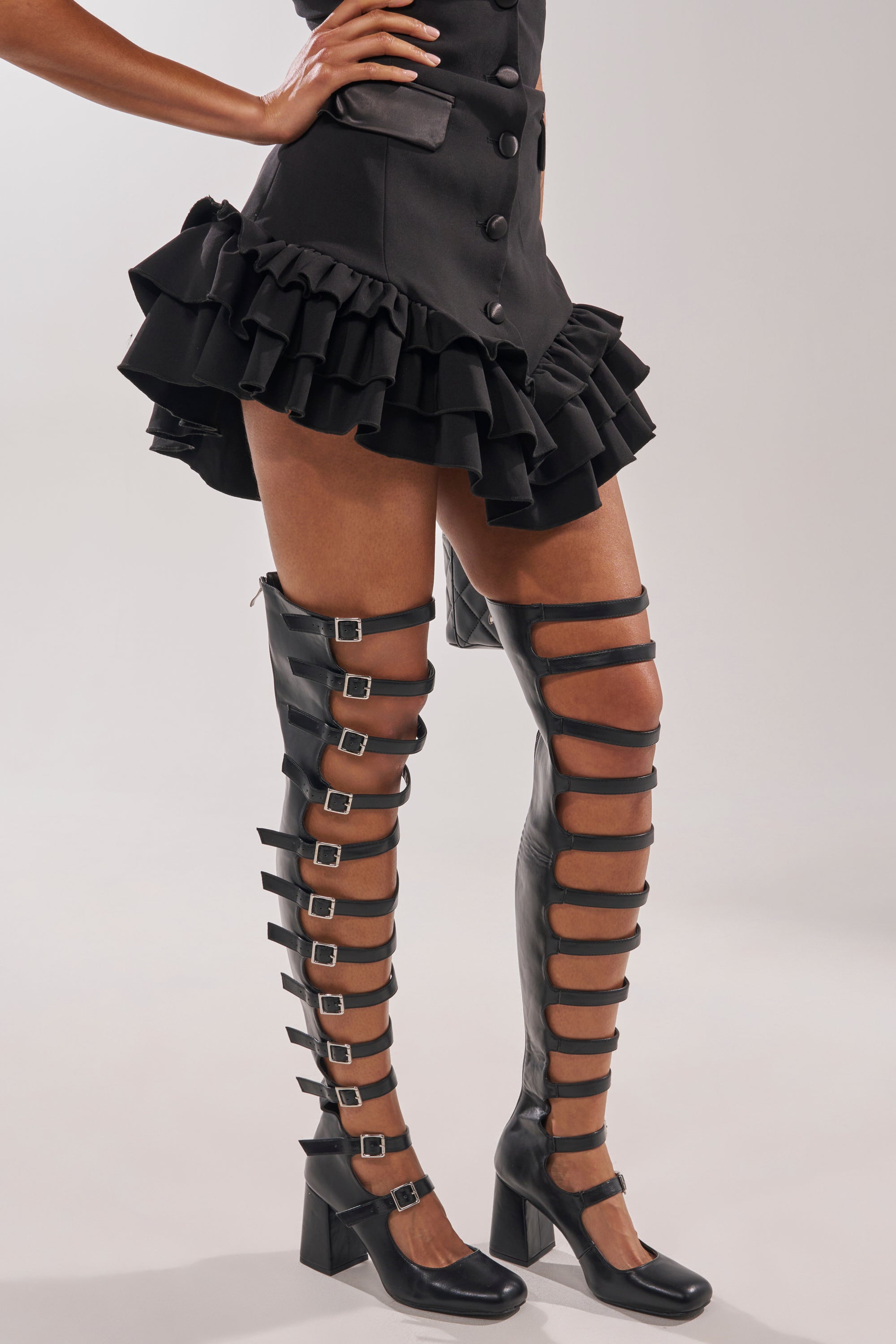 A person wearing a black ruffled mini skirt and the AKIRA GOLD YVETTE BLACK OVER THE KNEE MARY JANE BOOT, standing against a plain light background. 2