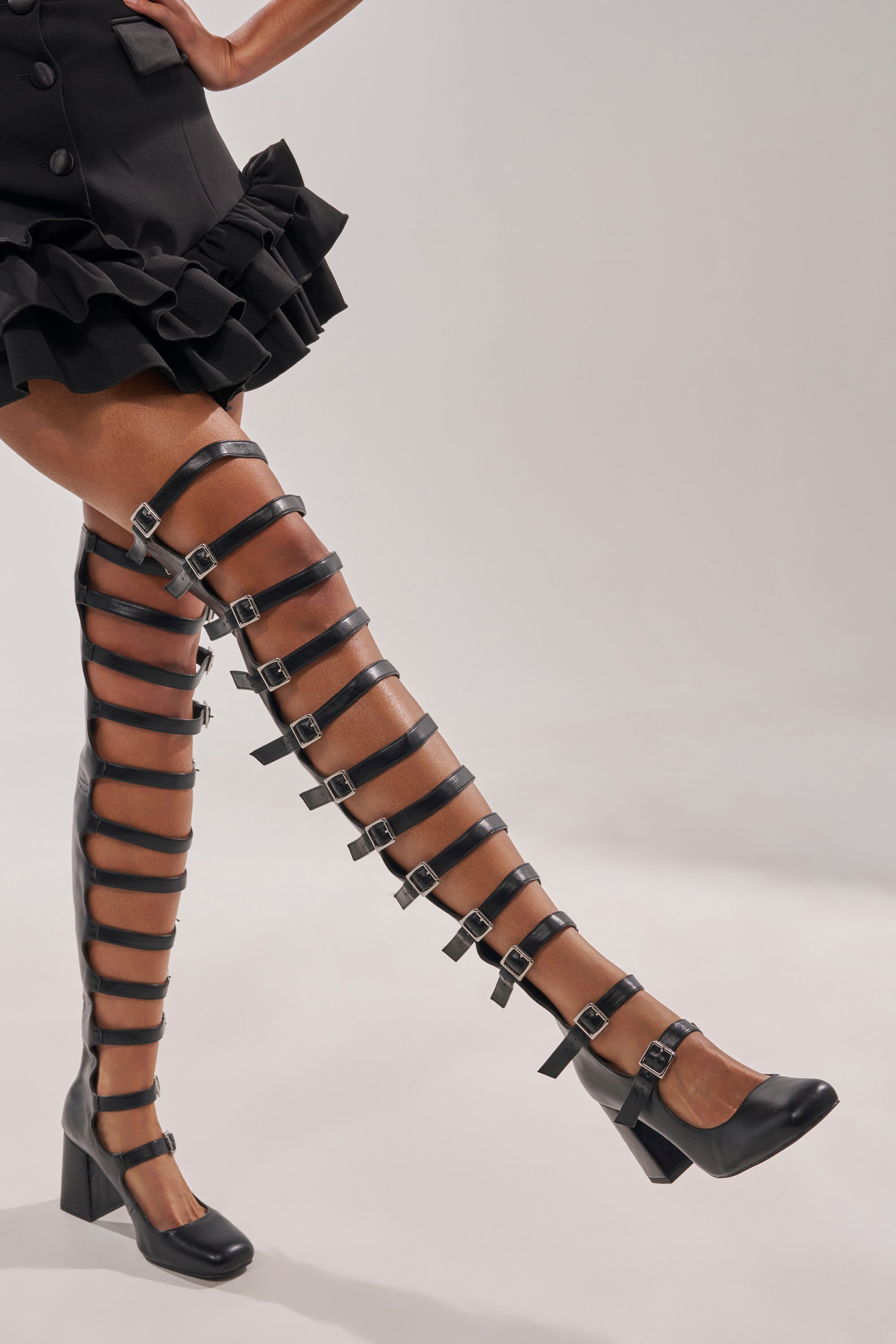 Someone poses against a plain light background wearing the AKIRA GOLD YVETTE BLACK OVER THE KNEE MARY JANE BOOT with sheer stockings and a black ruffled skirt. The boots feature faux leather, multiple silver buckles, and block heels. 3