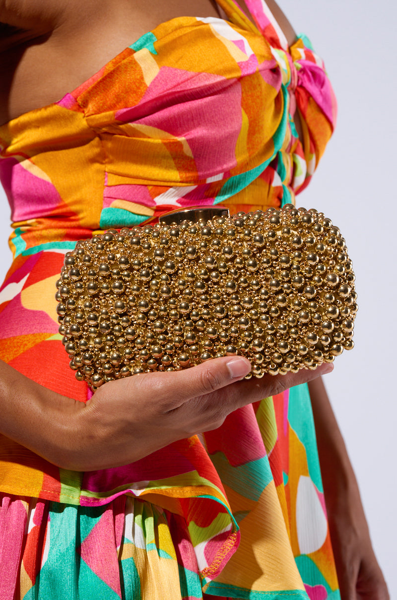 A person wearing a vibrant, multicolored dress holds a POPPIN BUBBLY EMBELLISHED CLUTCH in their left hand. The metallic gold hardshell clutch is adorned with numerous small, shiny golden beads, giving it a textured and luxurious appearance. It features a detachable chain link shoulder strap for versatile styling. 0