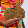 A person wearing a vibrant, multicolored dress holds a POPPIN BUBBLY EMBELLISHED CLUTCH in their left hand. The metallic gold hardshell clutch is adorned with numerous small, shiny golden beads, giving it a textured and luxurious appearance. It features a detachable chain link shoulder strap for versatile styling.