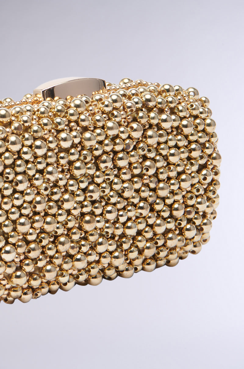 A close-up of the POPPIN BUBBLY EMBELLISHED CLUTCH showcases its luxurious and textured surface, adorned with numerous small gold metallic beads. The plain, softly lit background highlights the intricate details of the beadwork and the clutch's detachable chain link shoulder strap. 3