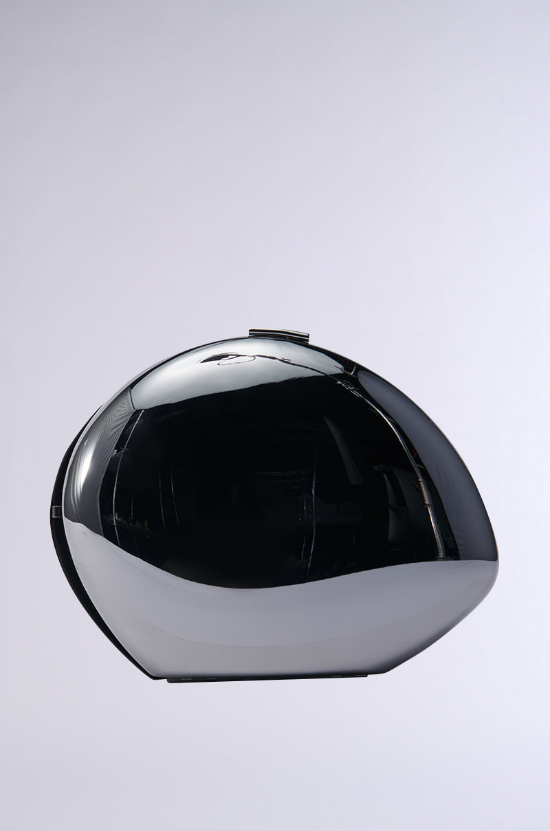 A sleek, metallic silver hardshell sculpture with a reflective black, oval-shaped design stands against a neutral gray background. Its surface is smooth and polished, embodying the essence of the MIDNIGHT DREAMIN METAL CLUTCH.