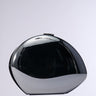 A sleek, metallic silver hardshell sculpture with a reflective black, oval-shaped design stands against a neutral gray background. Its surface is smooth and polished, embodying the essence of the MIDNIGHT DREAMIN METAL CLUTCH.