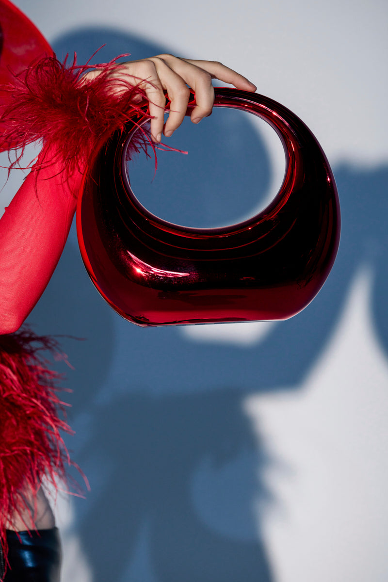 A person in a red feathered sleeve holds the CHERRY ON TOP CHROME PURSE, a shiny sculptural hardshell bag with a circular handle, against a light background that casts a dramatic shadow on the wall. 0
