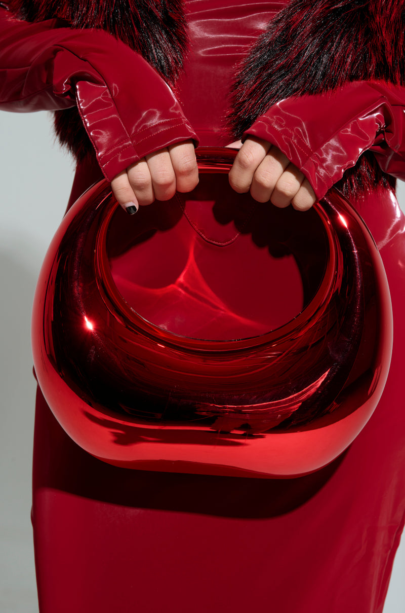 A person dressed in a glossy, red outfit with fur trim holds the CHERRY ON TOP CHROME PURSE. The plain gray background highlights the deep red hue and shiny chrome finish of the bag. 2