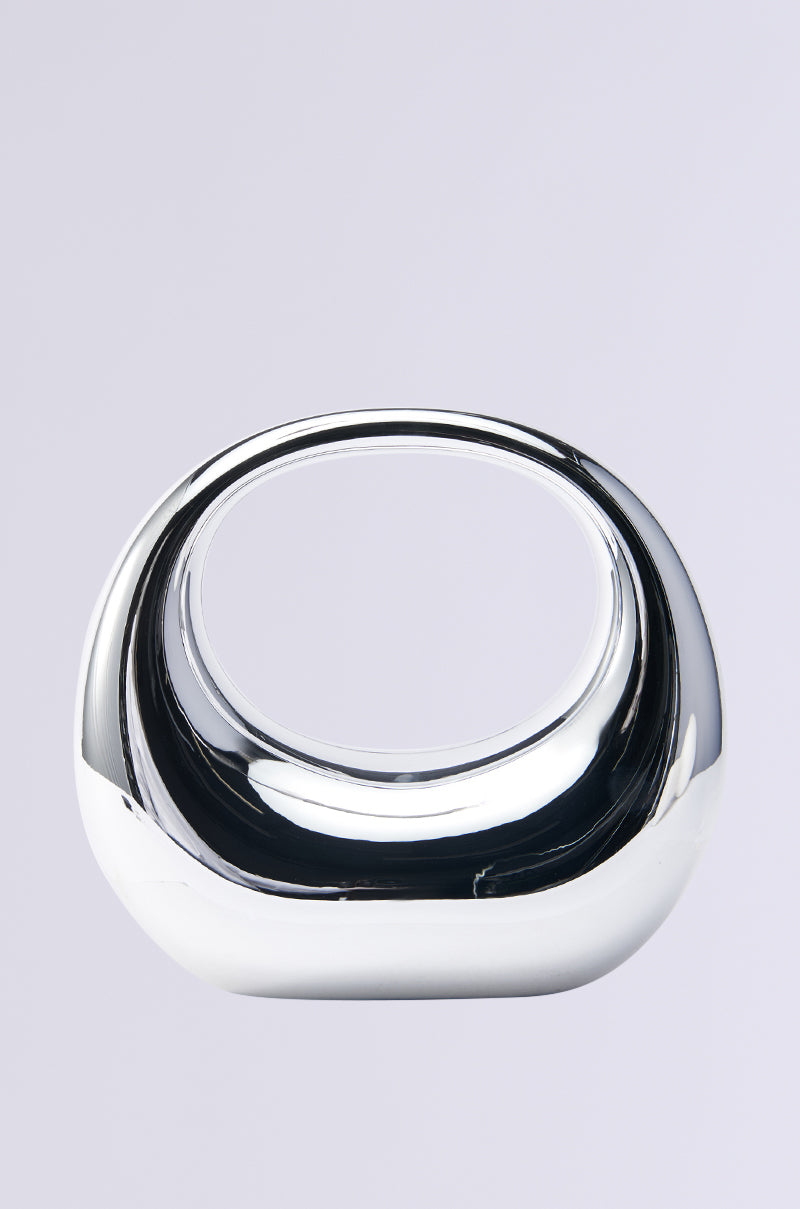 A modern, silver metallic sculpture with a smooth oval shape and open center set against a plain white background. Its reflective surface mirrors the sleek allure of the ALL I SEE IS CHROME TOP HANDLE PURSE, offering a contemporary and stylish look.