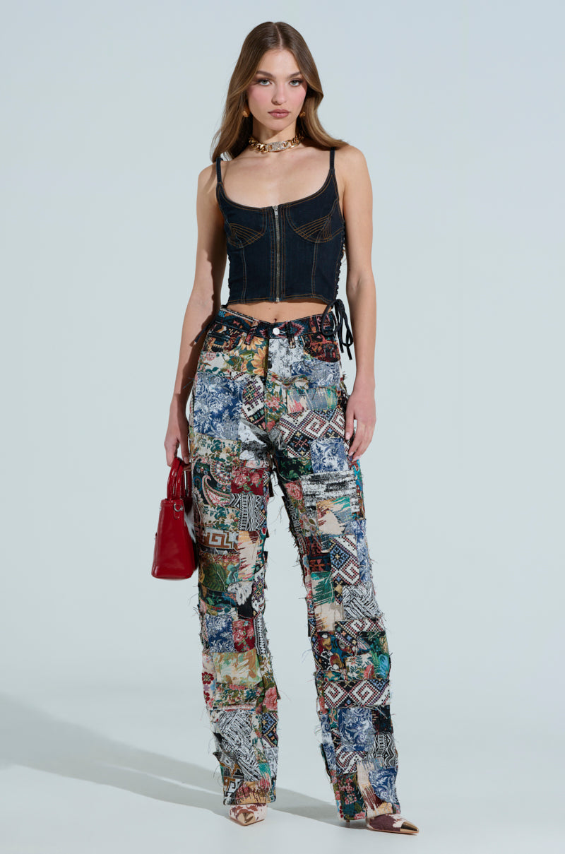 A woman with flowing long hair is wearing a corset-style black top paired with ODESSA RELAXED FIT PATCHWORK PANTS, and she holds a red handbag. Completed with sandals and a choker necklace, the patchwork pants stand out against the plain, neutral background.