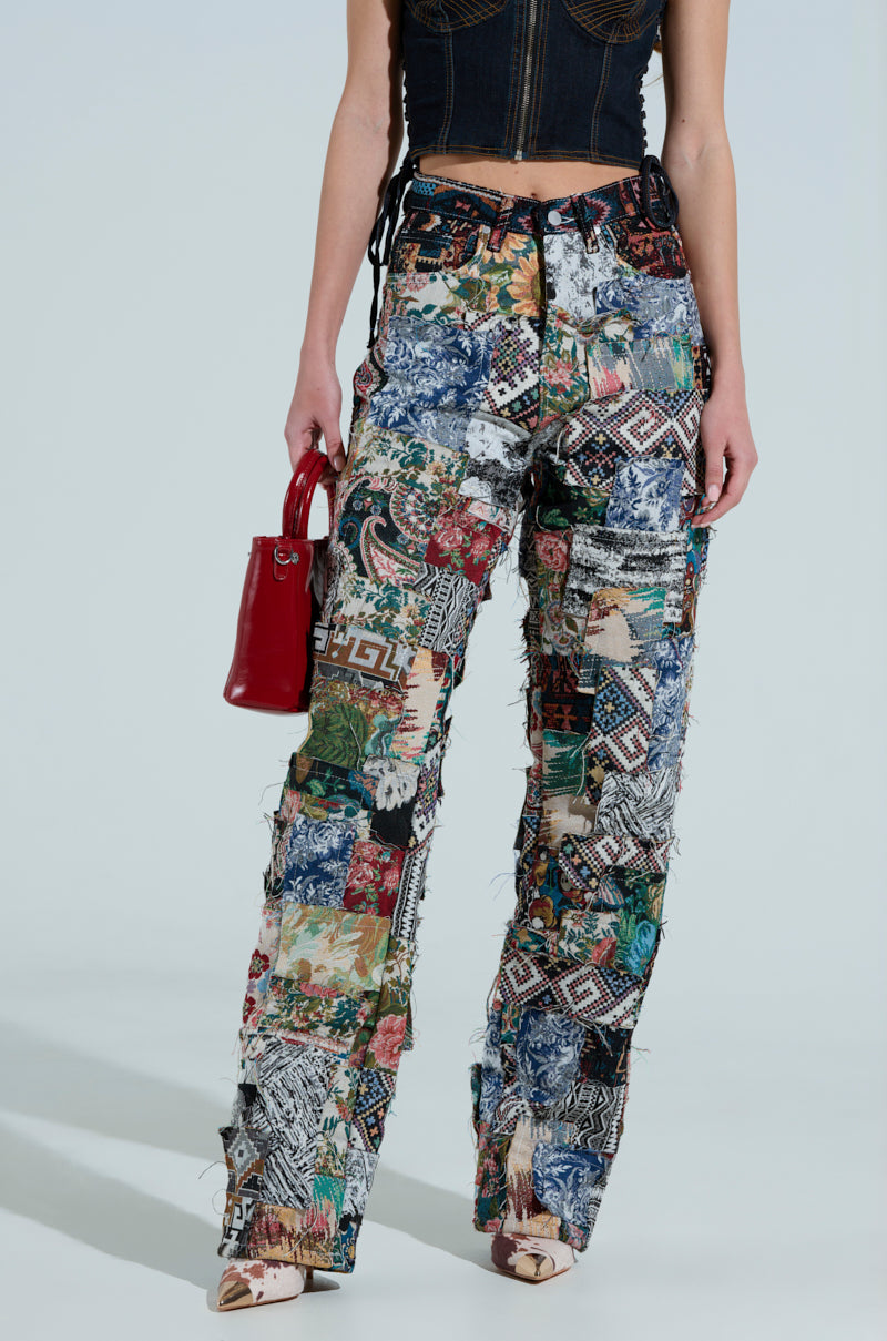 Someone shows off ODESSA RELAXED FIT PATCHWORK PANTS, made of tapestry-like material with colorful patterns, paired with a black sleeveless crop top and a red handbag, set against a plain, light-colored background.