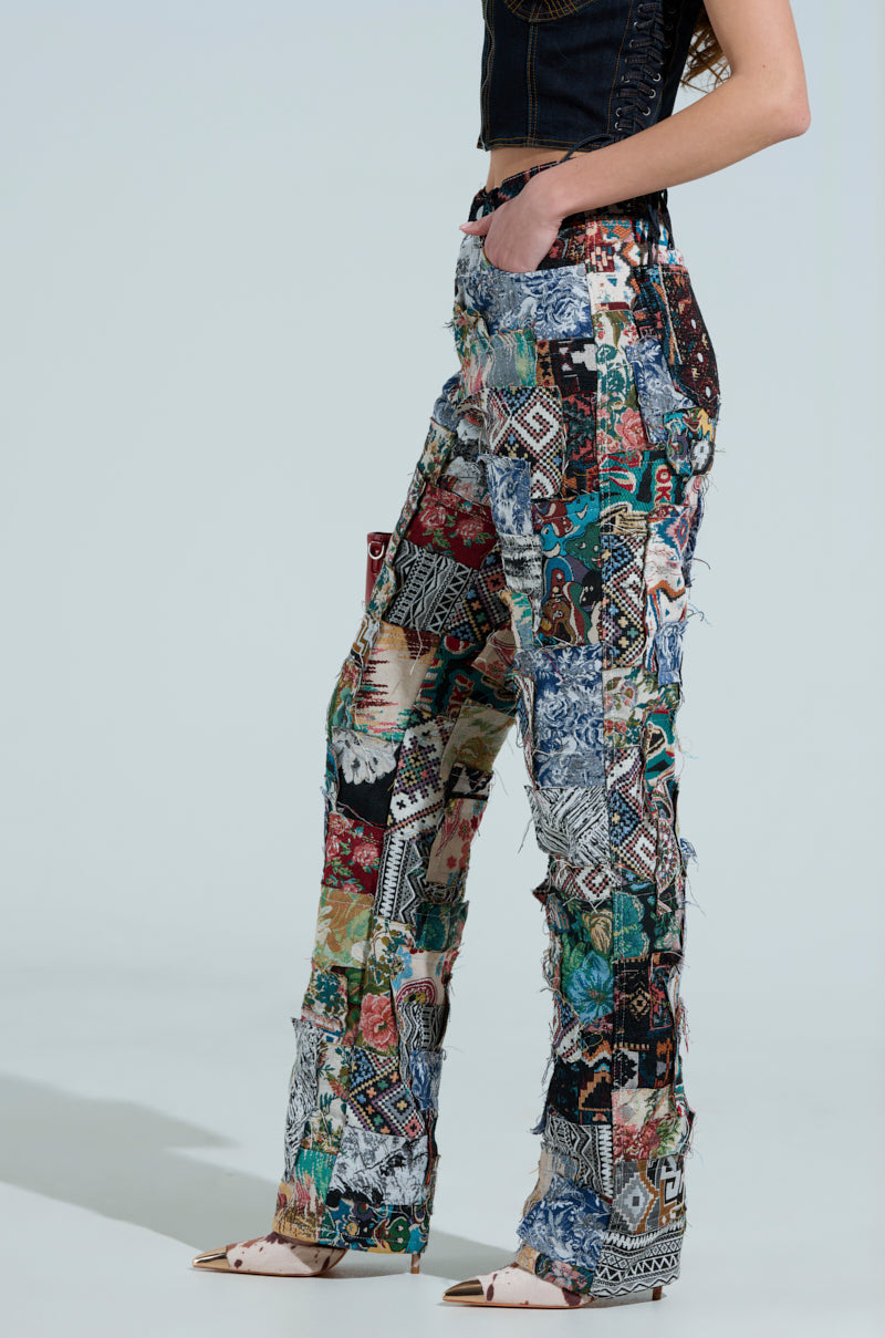Against a plain background, only the lower half of someone is visible as they wear high-waisted, flared Odessa Relaxed Fit Patchwork Pants with colorful tapestry-like material, paired with a sleeveless black top and pointed shoes.