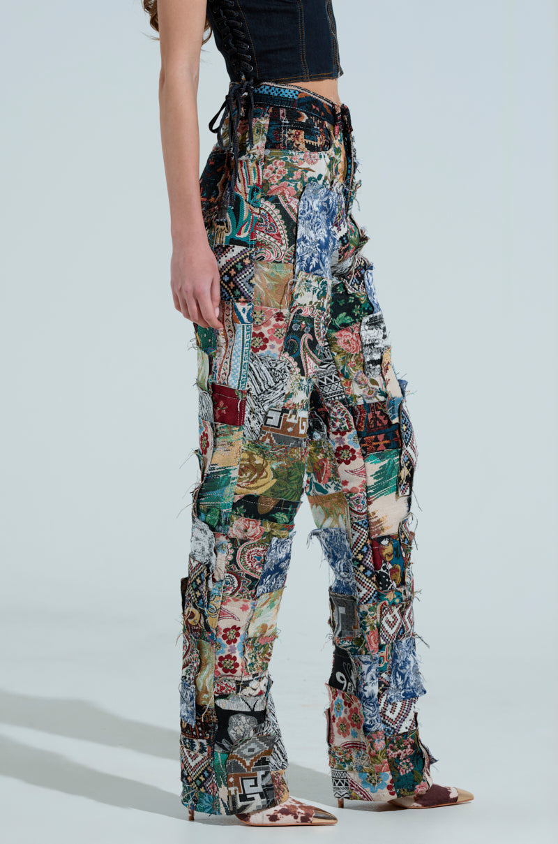 A person in ODESSA RELAXED FIT PATCHWORK PANTS, showcasing vibrant, tapestry-like patterns and textures in a side view, paired with a dark sleeveless top against a plain background.