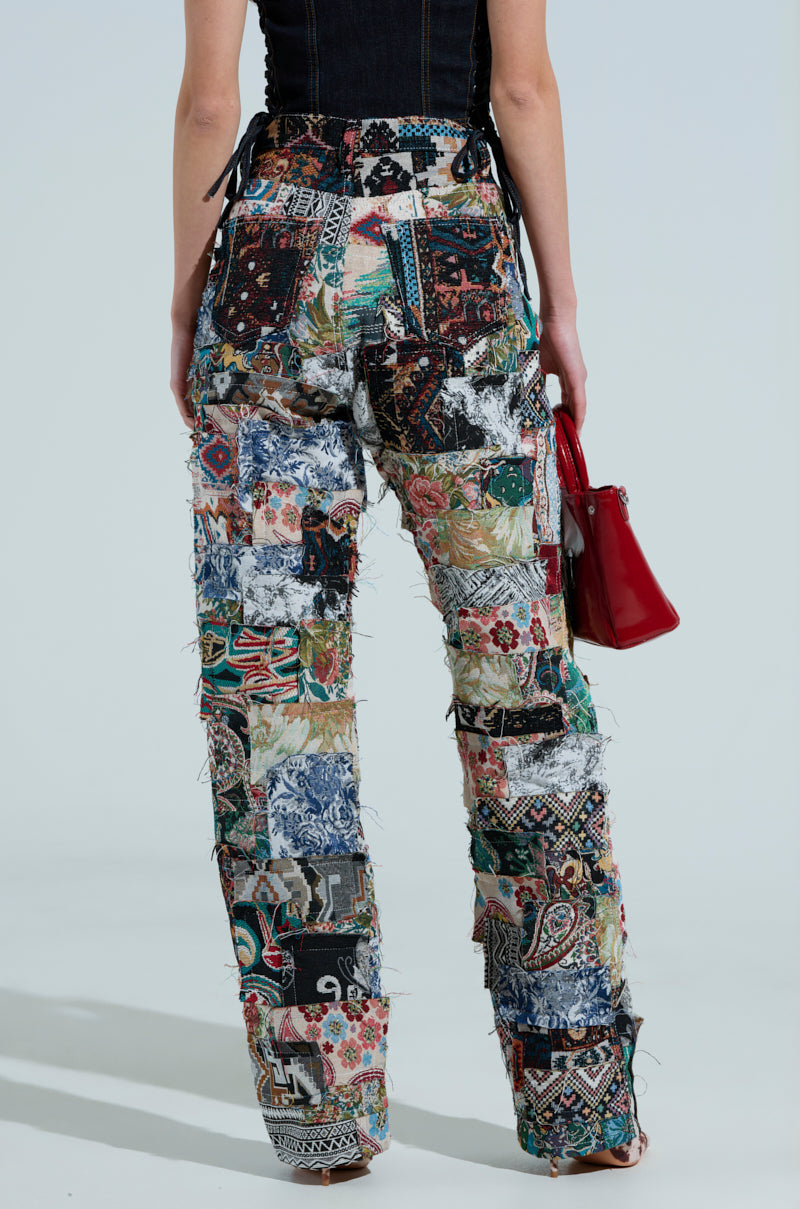 A person, back to the camera, is wearing Odessa Relaxed Fit Patchwork Pants made from tapestry-like material with vibrant patterns and holds a small red bag against a solid light background.