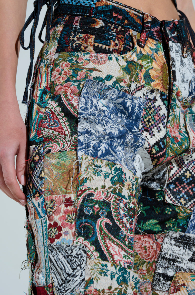 A close-up highlights a person wearing the ODESSA RELAXED FIT PATCHWORK PANTS, made from vibrant tapestry-like material with intricate floral, paisley, and abstract patterns. These designs blend bright and muted colors for a striking visual appeal.