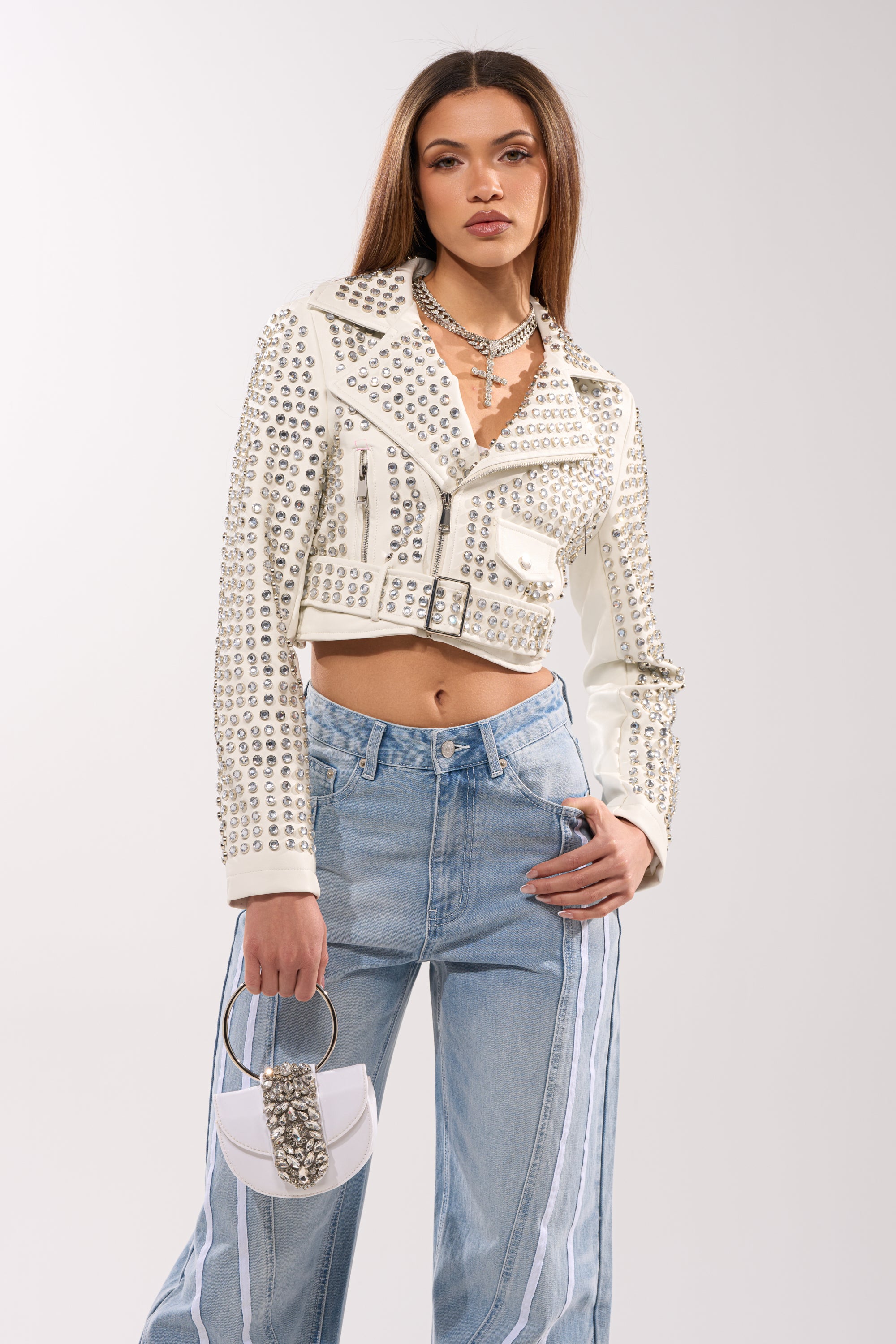 A woman wears the DEMI EMBELLISHED MOTO IN WHITE, paired with light blue high-waisted striped jeans, layered necklaces, and a small white handbag with metallic accents.