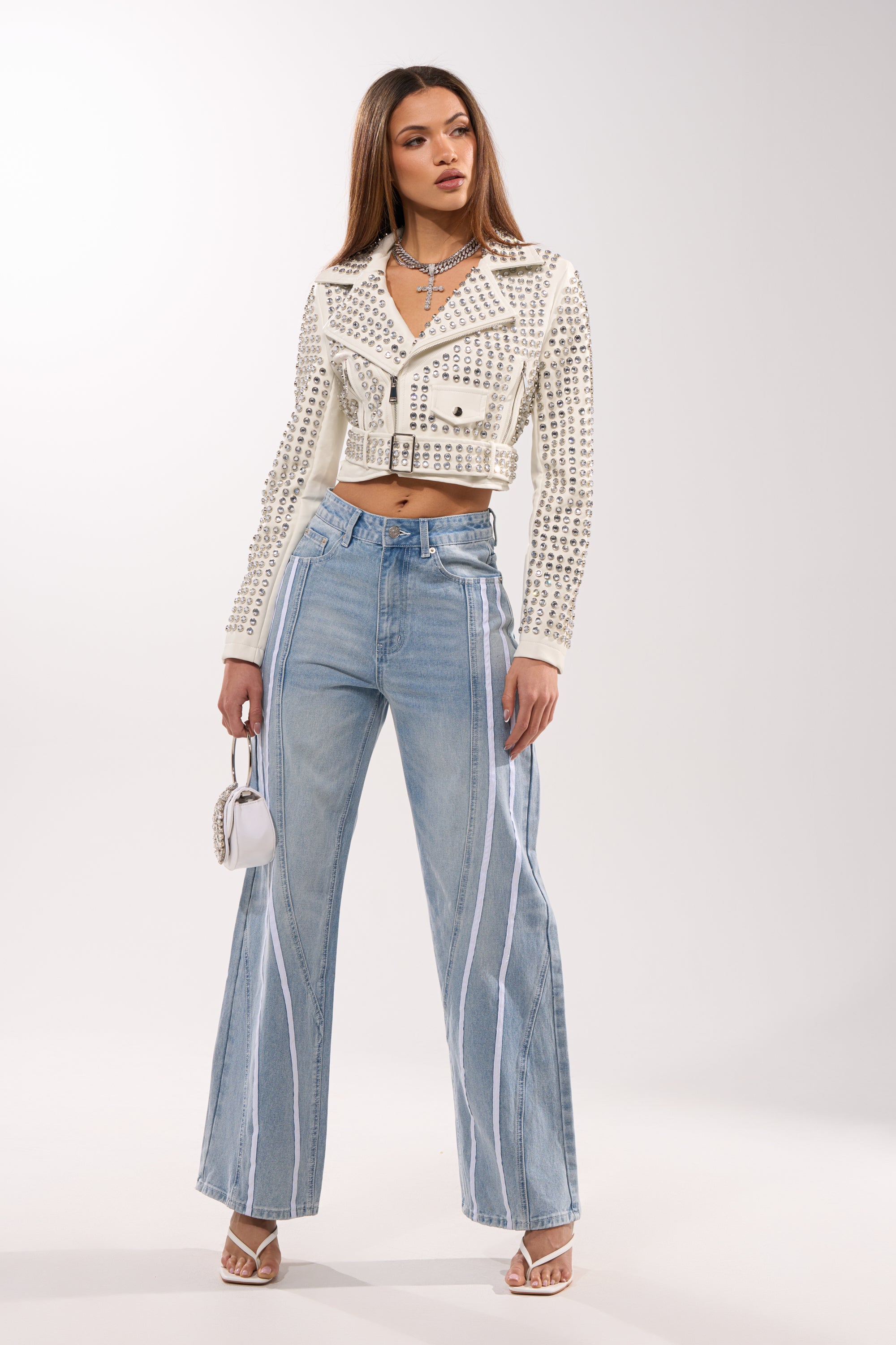 A woman poses in the DEMI EMBELLISHED MOTO IN WHITE, paired with high-waisted light blue wide-leg jeans with white stripes, white heels, and a small white handbag against a plain light background.