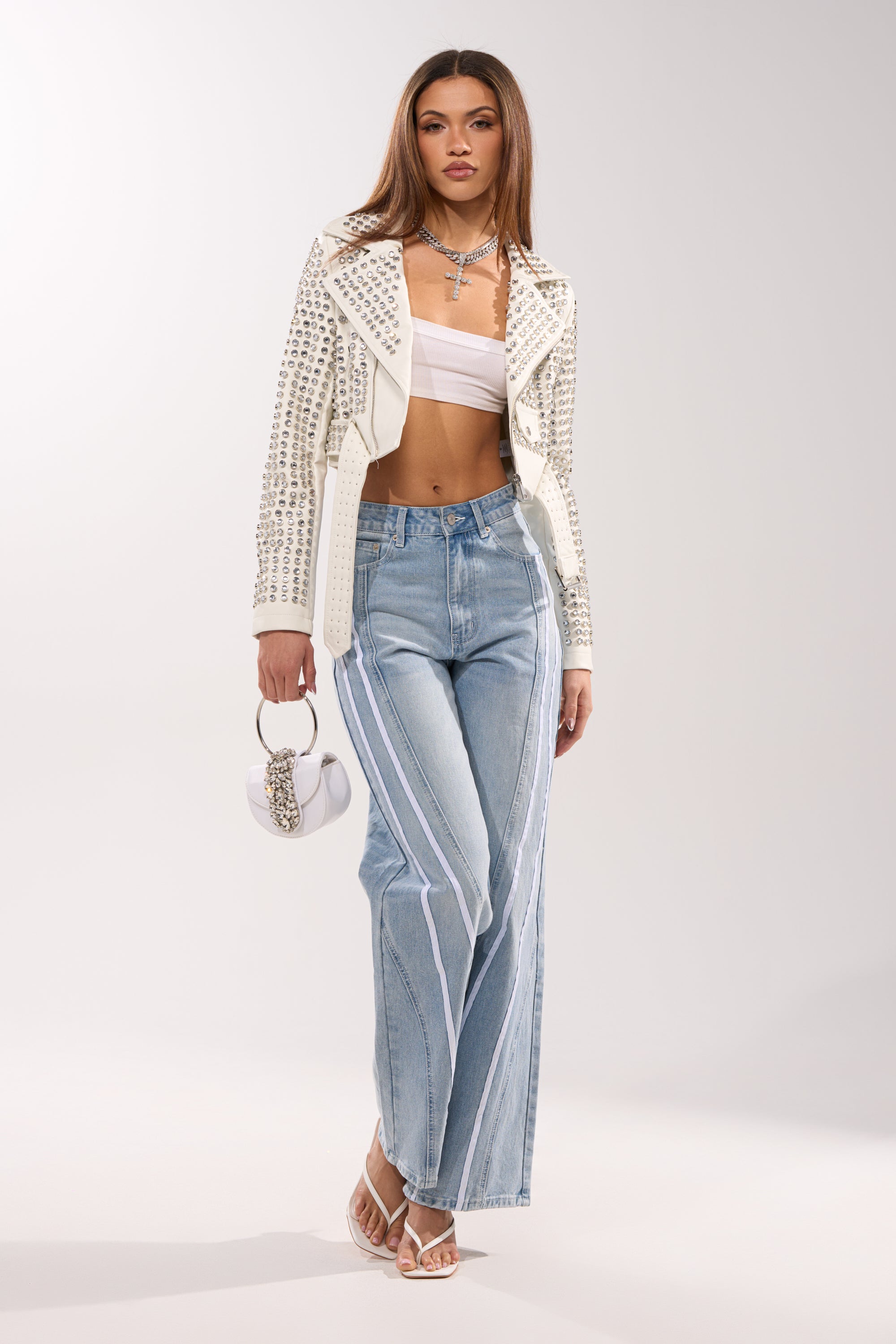 A woman stands confidently in the DEMI EMBELLISHED MOTO IN WHITE, paired with a white bandeau top, light blue wide-leg jeans with white stripes, white heeled sandals, layered necklaces, and a small white handbag.