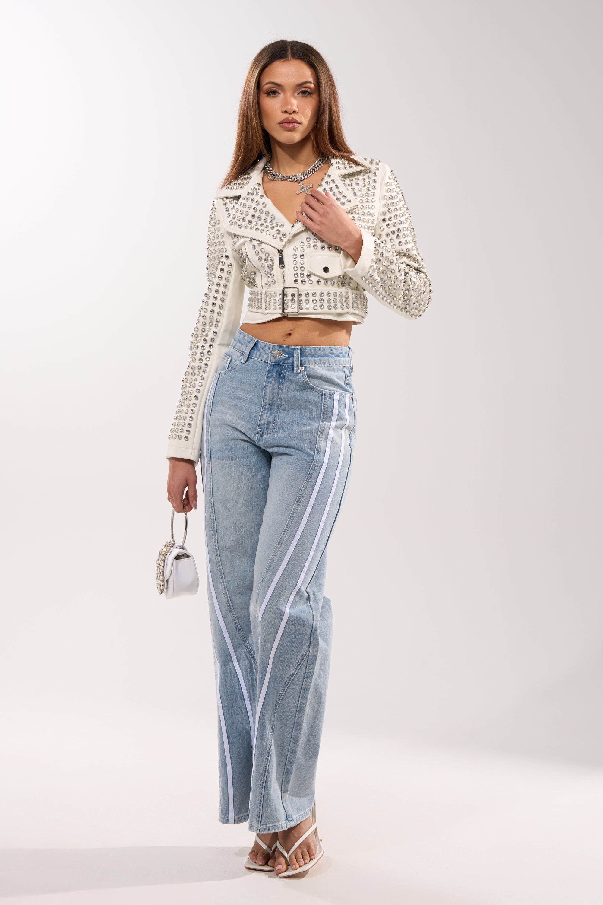 A woman stands confidently in the DEMI EMBELLISHED MOTO IN WHITE, light blue high-waisted flared jeans with white side stripes, silver heels, a silver necklace, and a small white purse against a plain background.