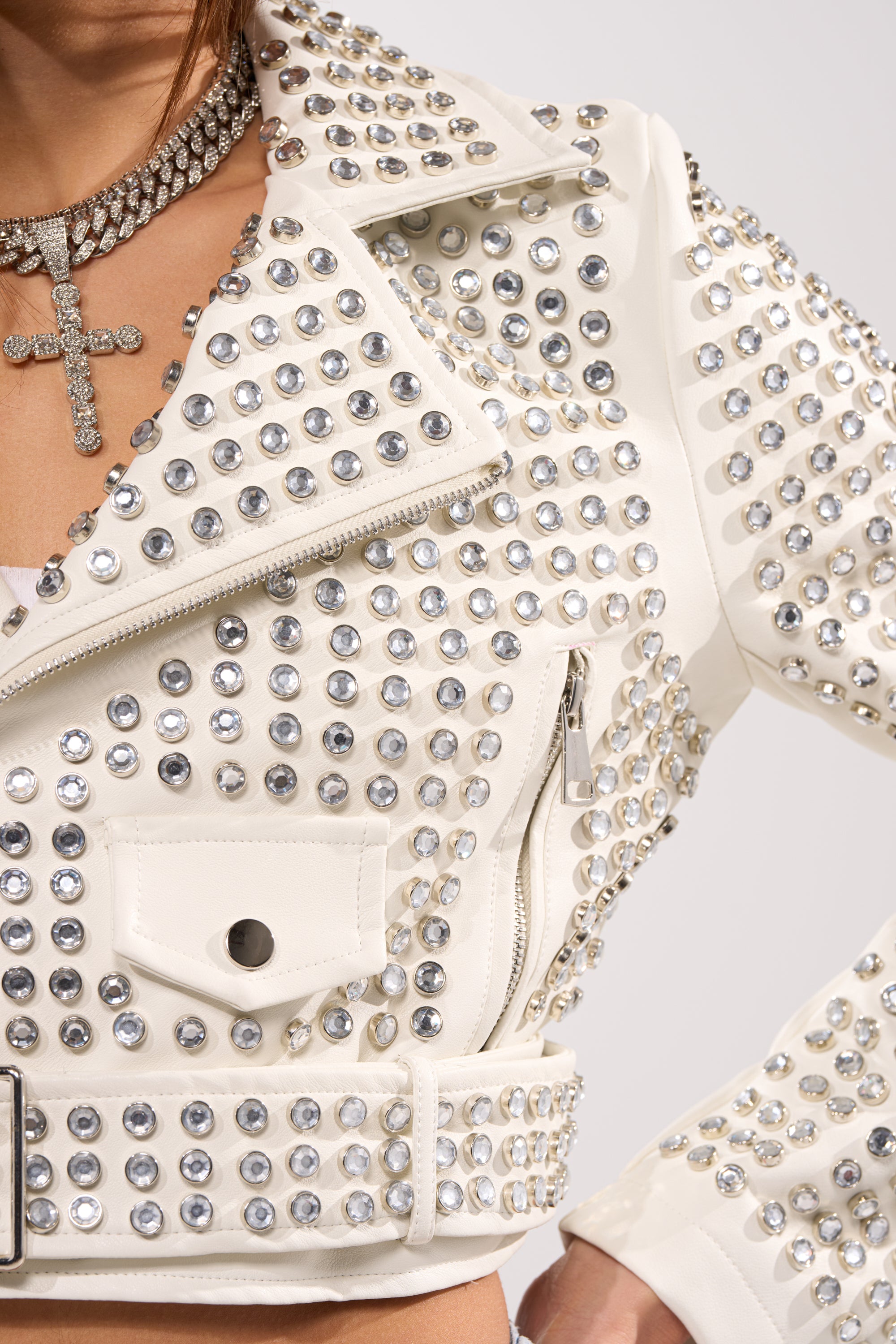 A close-up of the DEMI EMBELLISHED MOTO IN WHITE, adorned with silver rhinestones. It's styled with a rhinestone choker necklace featuring a large cross pendant. Only the model's lower face and upper body are visible.