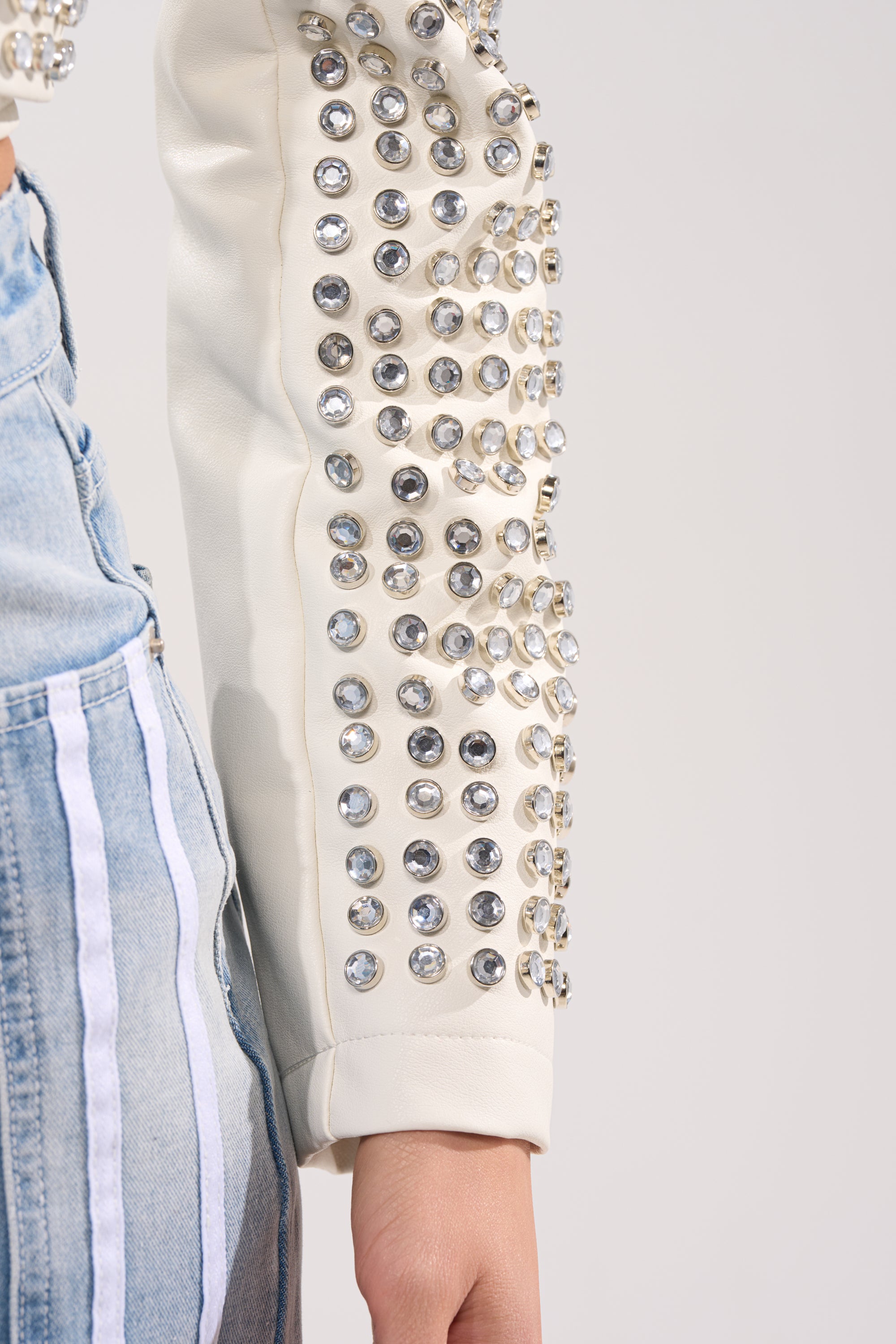 A close-up of a person wearing the DEMI EMBELLISHED MOTO IN WHITE, a cream-colored jacket with rows of round rhinestones on the sleeve, paired with light blue jeans featuring vertical white stripes.