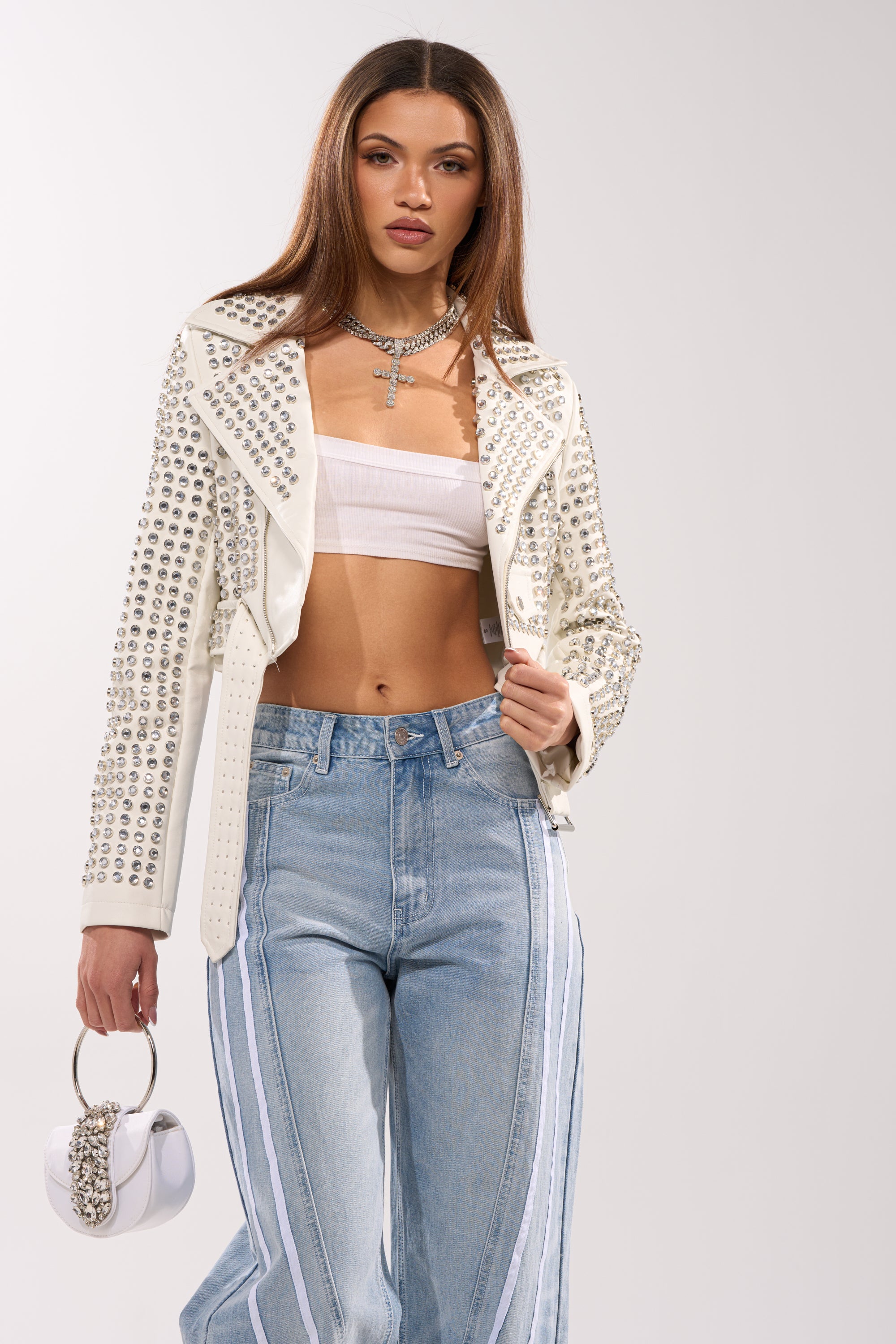 Wearing the DEMI EMBELLISHED MOTO IN WHITE over a white bandeau top and high-waisted light blue jeans with white side stripes, she holds a small white handbag and accessorizes with layered necklaces.