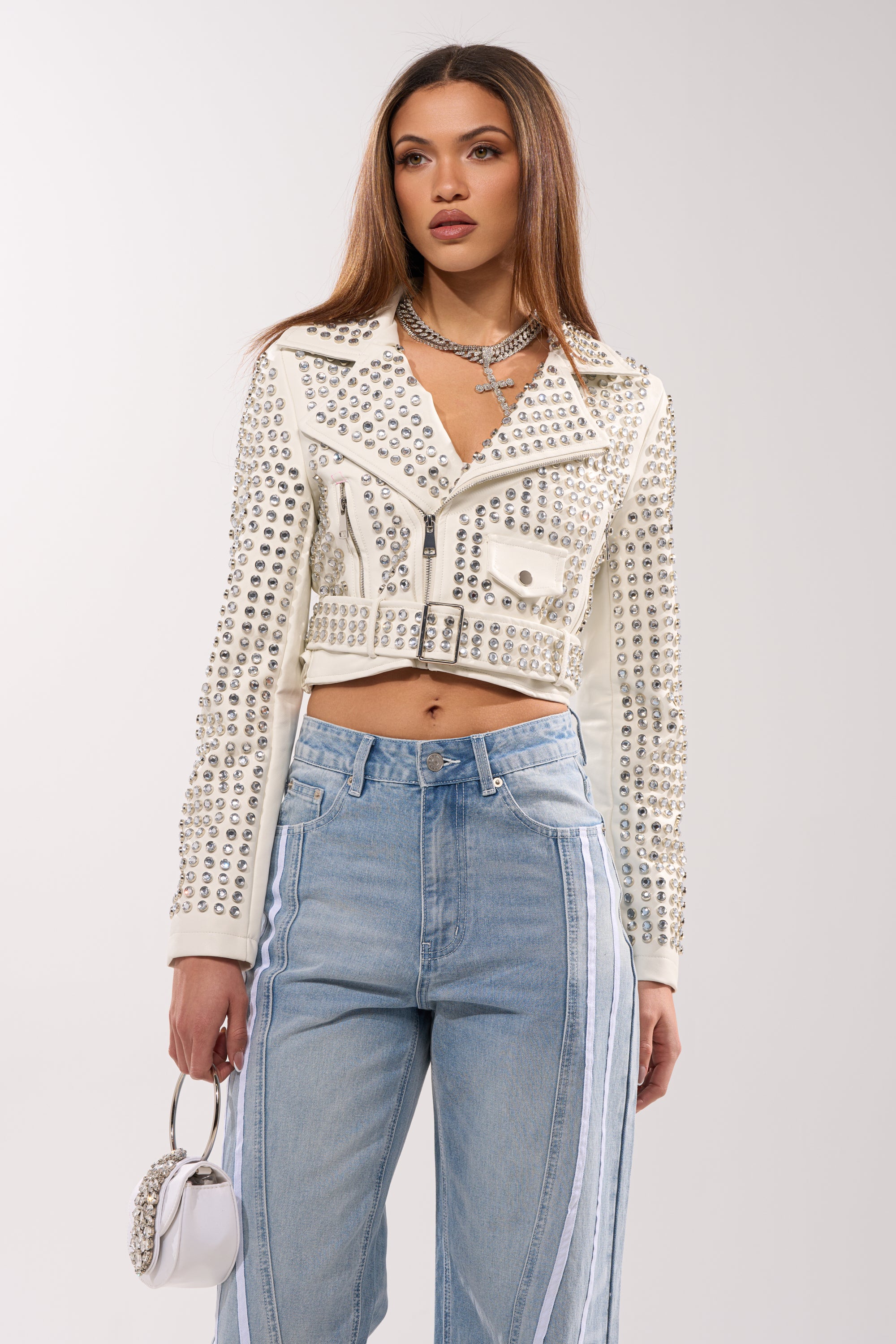 A woman wears the DEMI EMBELLISHED MOTO IN WHITE, styled with light blue high-waisted jeans with white side stripes, layered necklaces, and a small white handbag.