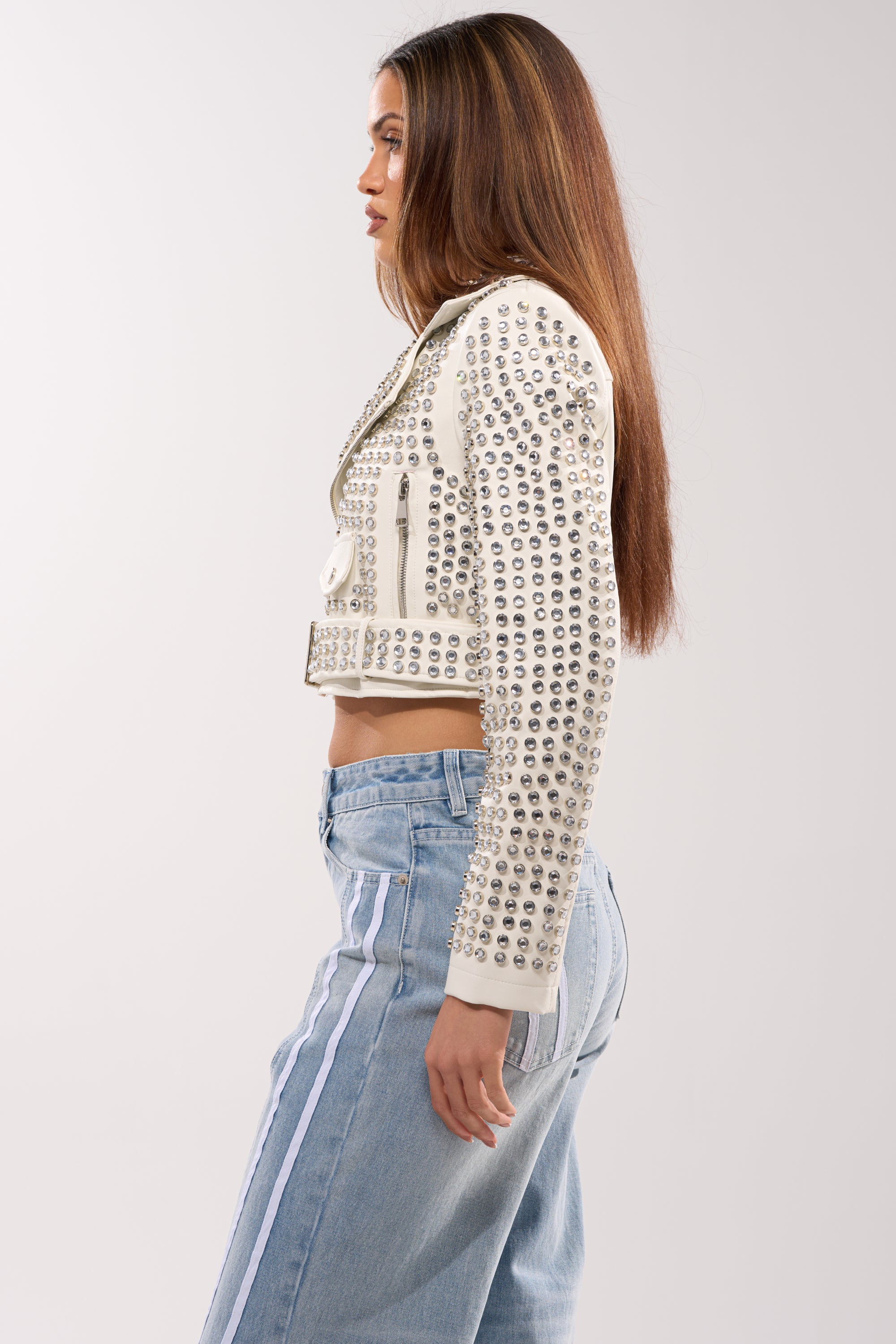 A woman with long brown hair stands in profile wearing the DEMI EMBELLISHED MOTO IN WHITE and light blue jeans with white side stripes, set against a plain light background.
