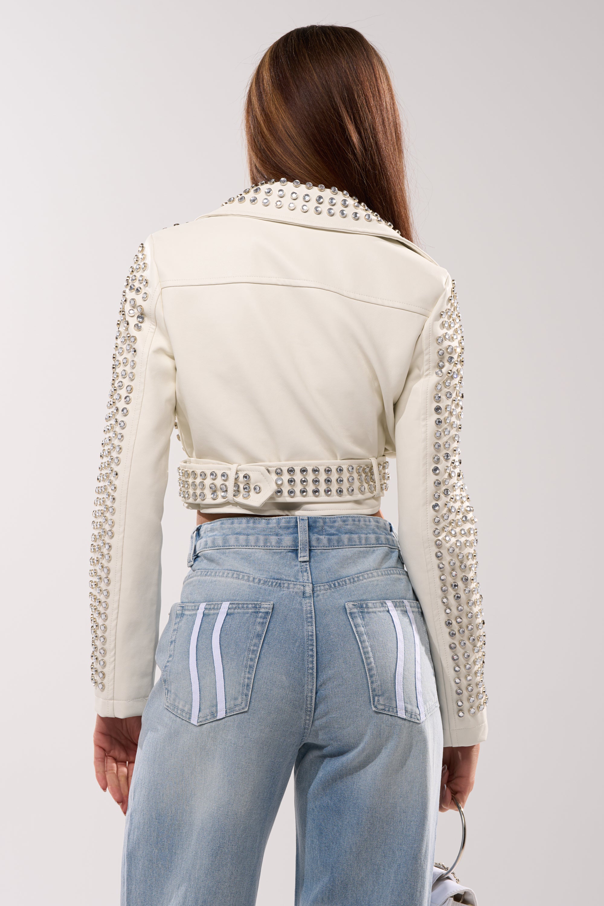 A woman with long brown hair is seen from behind wearing the DEMI EMBELLISHED MOTO IN WHITE and light blue jeans with white pocket stripes, holding a small handbag in her left hand.
