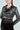 Wearing the DEMI EMBELLISHED MOTO, a person in a black faux leather jacket with silver studs and a belted waist stands arms crossed. The jacket complements their sheer lace dress, long hair, and large hoop earrings.