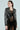 A woman with dark hair tied back wears the DEMI EMBELLISHED MOTO jacket and a black lace dress. Hoop earrings complete her look as she gazes to the side against a plain background.